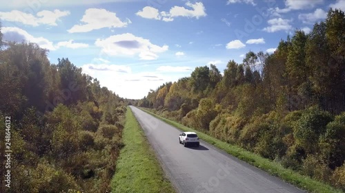 camera follows modern car driving along road among old wood against blue sky with light clouds