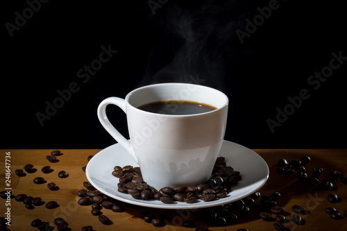 Brewed coffee in a white cup with steam or vapor and coffee beans scattered on a wooden surface and on the white saucer. 