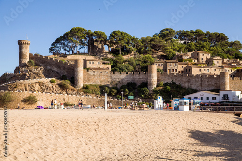 Beach under the castle in Tossa de Mar. People on the seafront near the walls of the old city. The historic city and the castle tower of La Vila Vella. A beautiful Catalan town on the Costa Brava