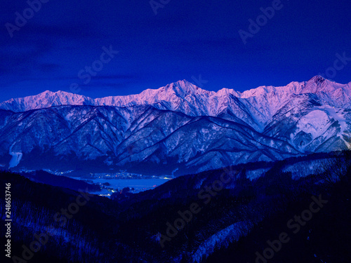 峠からのアルプス山脈を見渡す、夜明け前のブルー、谷間から町並みの街灯り、アルプス山脈は雪景色で朝焼前のピンク色に染る。