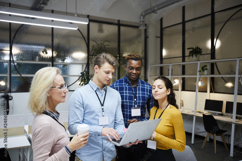 Group of multi-ethnic business colleagues standing in modern office and using portable computer while discussing internet project