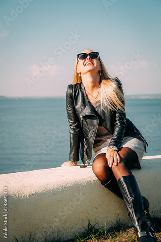 Blonde with long hair in a leather black jacket posing on the shore of the blue sea photo