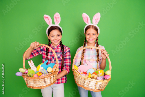 Portrait of two lovely sweet cheerful cheery positive pre-teen girls wearing checked shirt having fun holding in hands festive baskets isolated over bright vivid shine green background