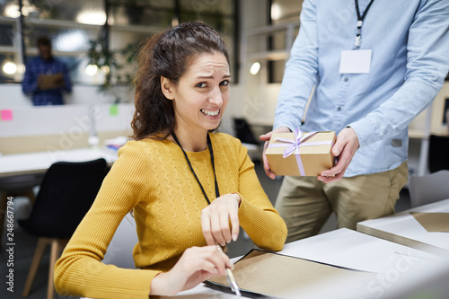 Displeased frowning attractive young lady in sweater disgusted by colleagues gift sitting at table and looking at camera while working on project design