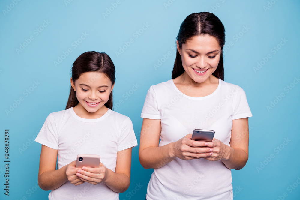 Close up photo beautiful two people brown haired mom small little daughter addicted telephones reader write sms got good news information wear white t-shirts isolated bright blue background Close up photo beautiful two people brown haired mom small little daughter addicted telephones reader write sms got good news information wear white t-shirts isolated bright blue background
