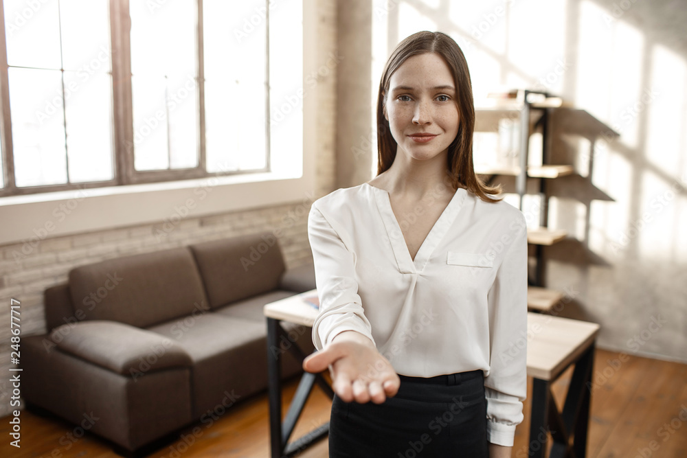 Young businesswoman reach hand to camera. She look straight and smile a bit. Empty room. Alone. Daylight. Young businesswoman reach hand to camera. She look straight and smile a bit. Empty room. Alone. Daylight.
