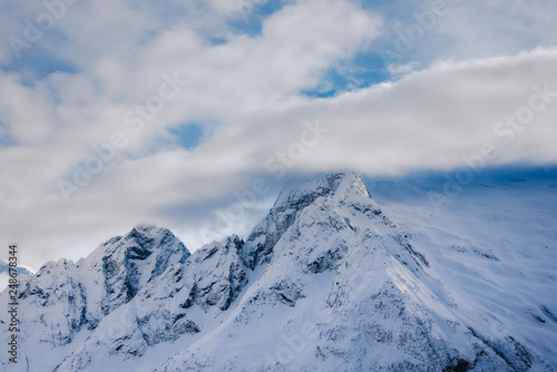mountain peaks of Dombai mountains covered with snow surrounded by thick clouds against the blue sky © Ambartsumian