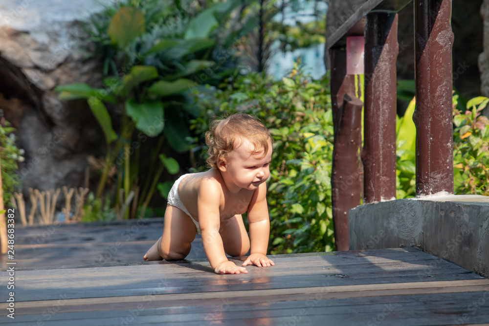Child crawls along the dark brown wooden walkway of the bridge at sunset. Baby learn crawl on the background of greenery and a wooden bridge. There is a place for text. Child crawls along the dark brown wooden walkway of the bridge at sunset. Baby learn crawl on the background of greenery and a wooden bridge. There is a place for text.