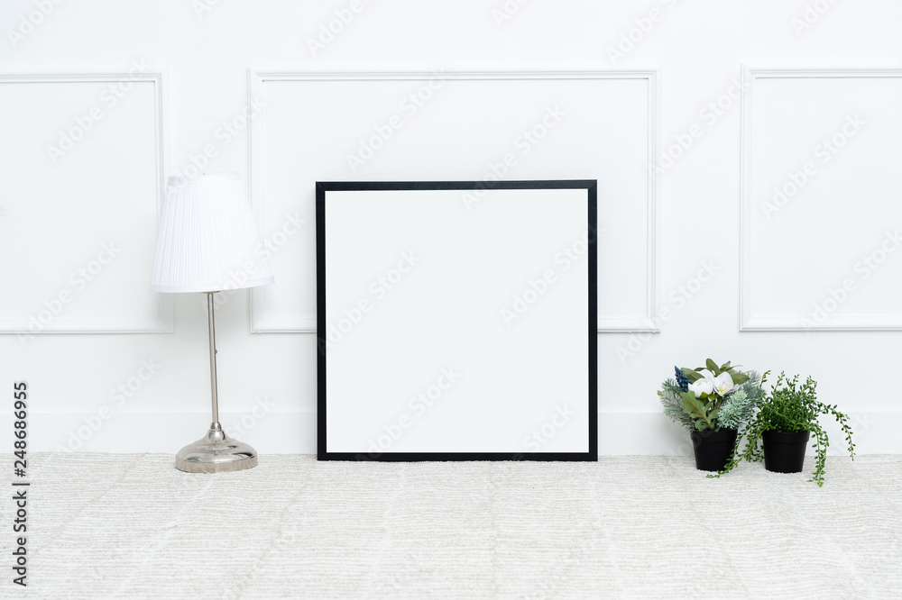There are beautiful flowers and various objects near the rectangle blank picture frame in front of white wall on the carpet living room.