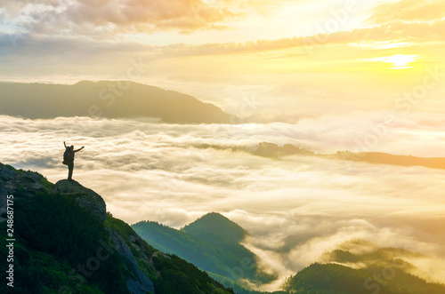 Wide mountain panorama. Small silhouette of tourist with backpack on rocky mountain slope with raised hands over valley covered with white puffy clouds. Beauty of nature  tourism and traveling concept
