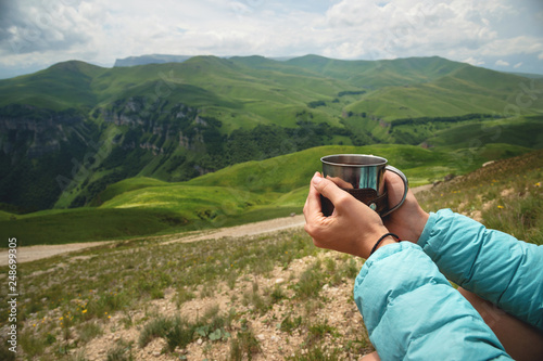 First-person view of a girl's hands holding a plastic cup of tea against a plateau of green hills and a cloudy sky in summer © yanik88