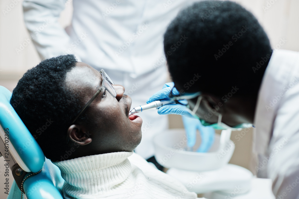 Fototapeta premium African american man patient in dental chair. Dentist office and doctor practice concept. Professional dentist helping his patient at dentistry medical. Drilling patient's teeth in clinic.