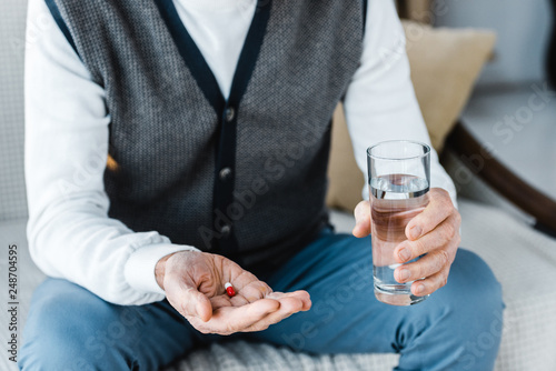cropped view of retired man holding pill and glass of water at home
