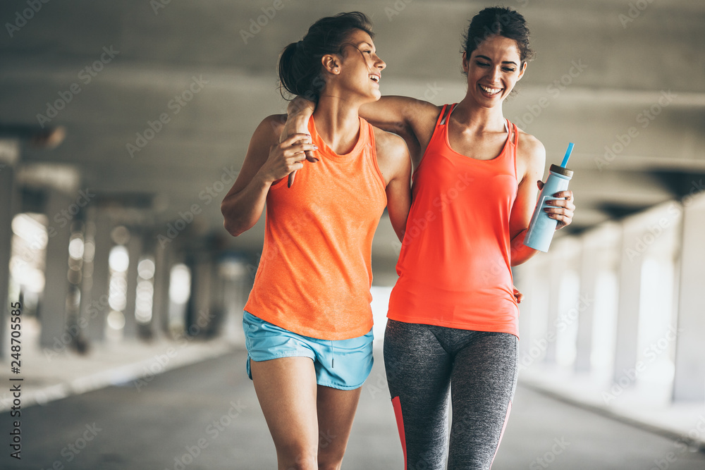 Two female friends jogging on the city street under the city road overpass.They relaxing after jogging and making fun.Embracing each other. - obrazy, fototapety, plakaty 