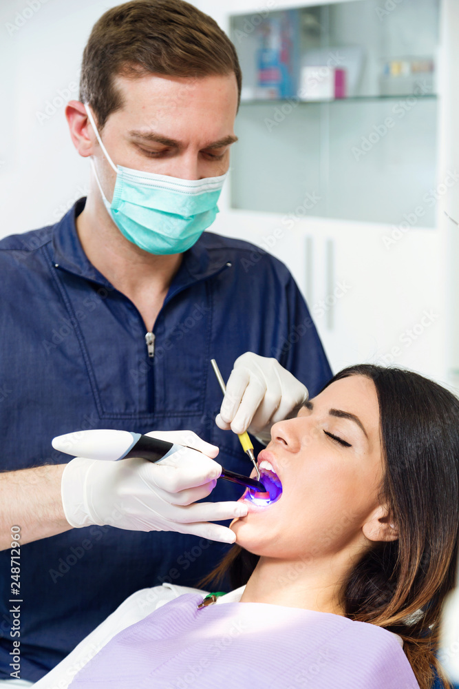 Handsome confident dentist whitening tooth to female patient on dental chair in dental clinic.