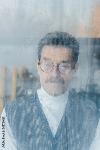 sad senior man in glasses standing at home near window