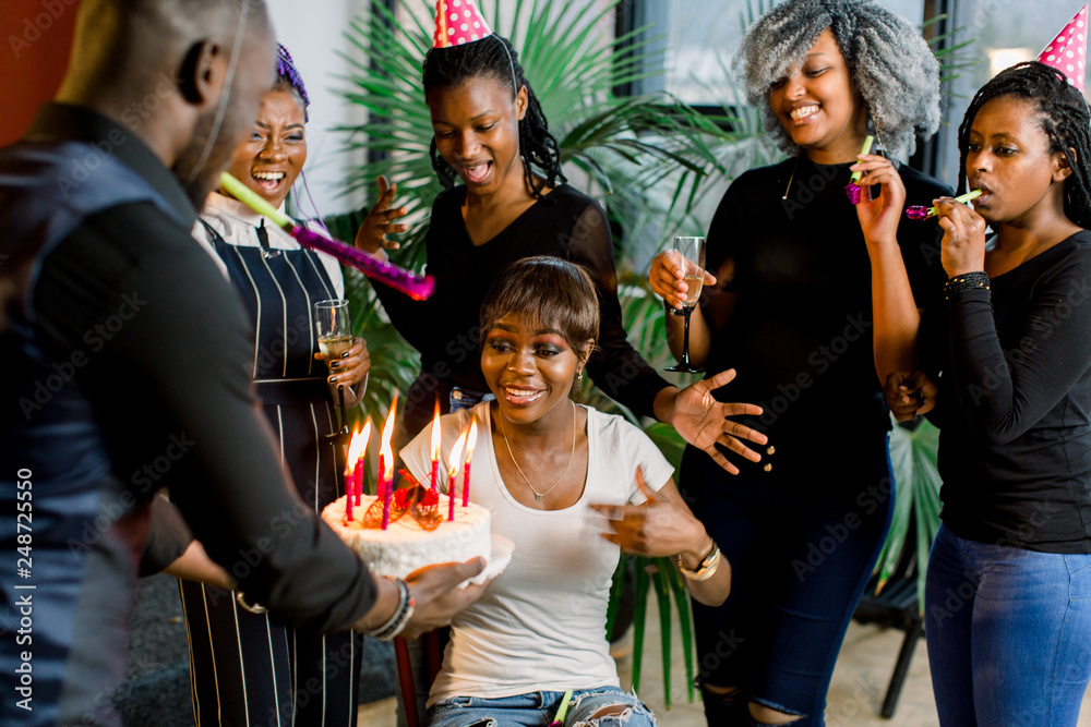 Fototapeta premium Portrait of charming African American girl blowing on candles on birthday cake surrounded by friends at party
