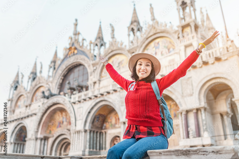 Young happy asian girl traveler with hat and backpack sitting on a parapet on San Marco square in Venice. Vacation and holidays in Italy and Europe concept