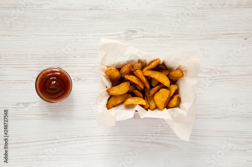 Fried potato wedges in paper box and barbecue sauce on a white wooden surface. From above, overhead, top view. Close-up.