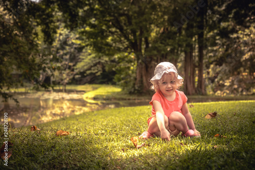 Beautiful smiling kid girl sitting alone on grass in lush green summer park in tree shadow childhood lifestyle