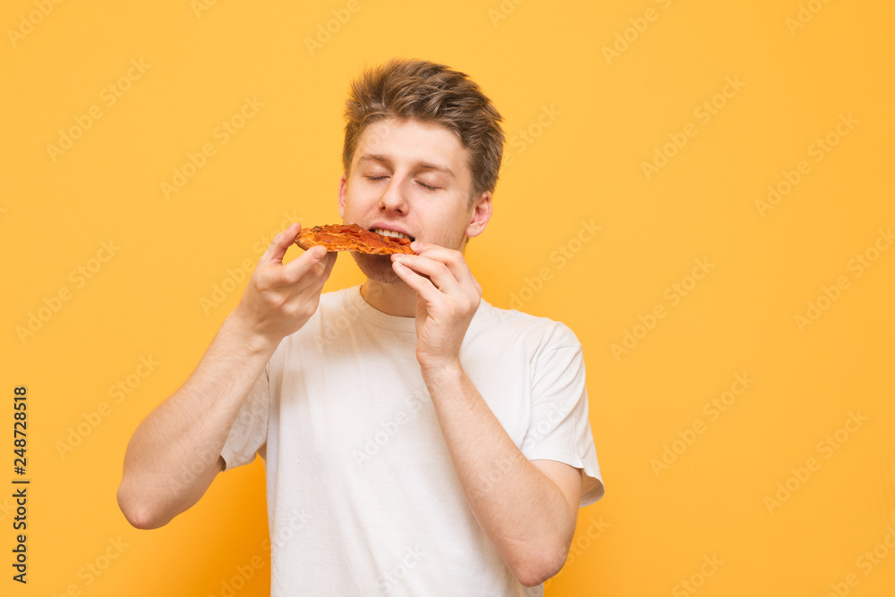 Young man eats a piece of appetizing pizza on a yellow background. Hungry guy holds a piece of pizza in his hands, looks at him and is going to eat it, isolated on a yellow Young man eats a piece of appetizing pizza on a yellow background. Hungry guy holds a piece of pizza in his hands, looks at him and is going to eat it, isolated on a yellow