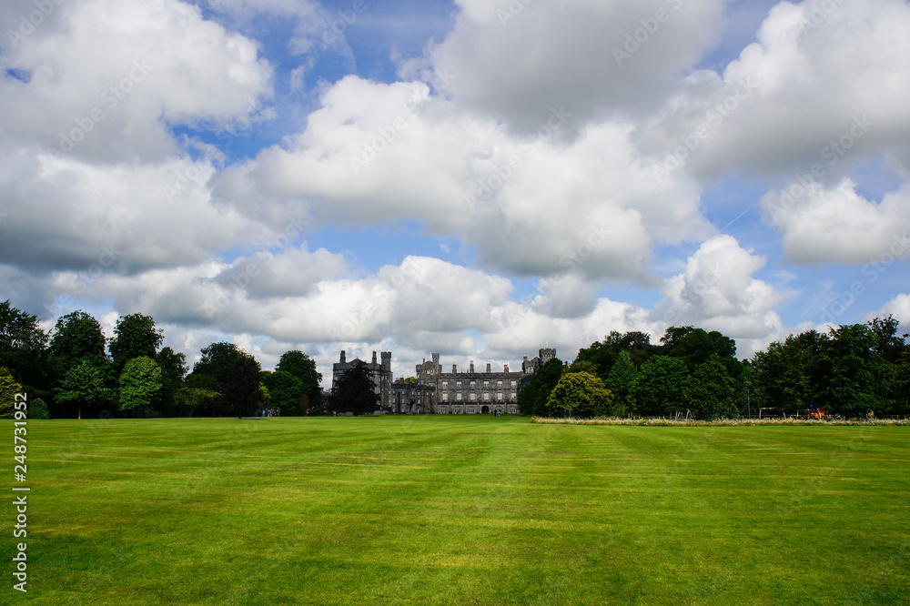 Distant castle in the forest under a beautiful blue sky and a vast green grass garden. Kilkenny Castle in Ireland.