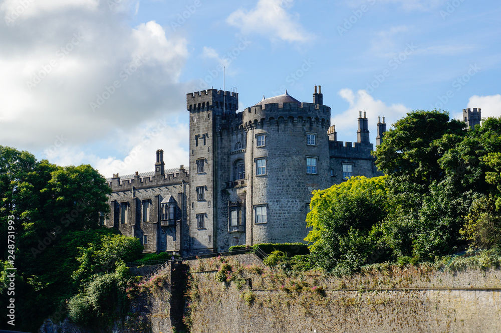 Fortified castle with grey stone walls surrounded by green trees under a blue sky with white clouds. Kilkenny Castle in Ireland.