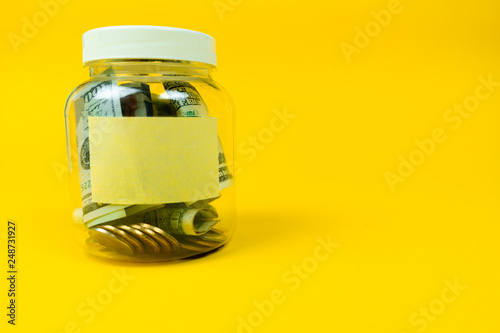 Money jar with coins and paper dollars with sticker for your text, copy space. Isolated on yellow background. Close up