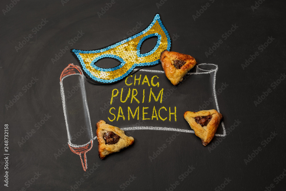 Painted scroll with an inscription Happy Purim on a chalk board next to the carnival mask and Hamantaschen cookies