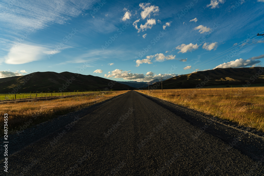 straight street during sunset with corn field on each side and mountains and blue sky in the background