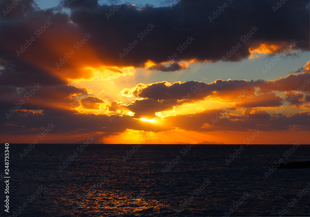 sunset over a calm dark sea with orange rays shining though dark dramatic evening clouds