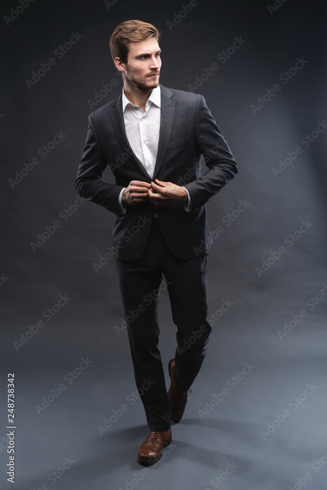 Full length portrait of a handsome young businessman dressed in suit isolated over gray background, posing, looking away, walking Full length portrait of a handsome young businessman dressed in suit isolated over gray background, posing, looking away, walking