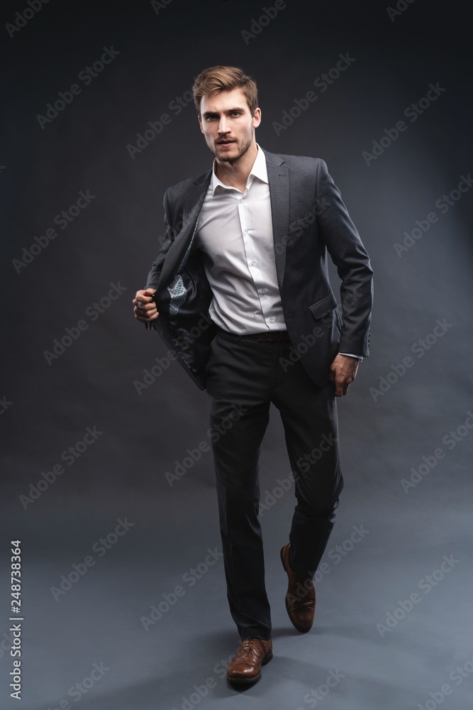 Full length portrait of a handsome young businessman dressed in suit isolated over gray background, posing, looking away, walking Full length portrait of a handsome young businessman dressed in suit isolated over gray background, posing, looking away, walking