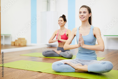 Portrait of two smiling young women sitting in lotus position during yoga workout in fitness club, copy space