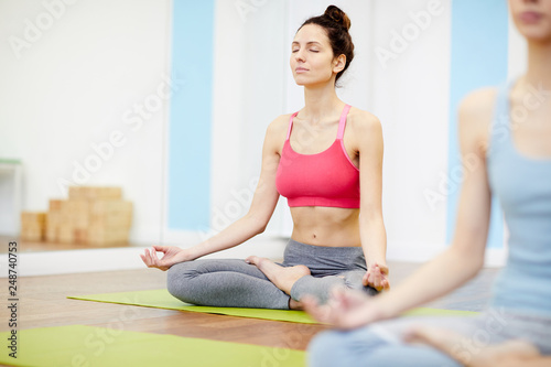 Fototapeta Naklejka Na Ścianę i Meble -  Full length portrait of smiling woman sitting in lotus position with eyes closed during yoga workout in fitness club, copy space