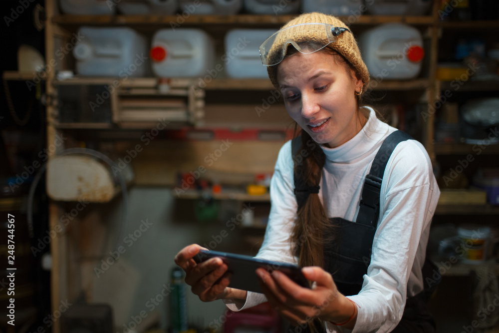 Portrait of young builder girl standing in workshop use smartphone, wearing construction glasses. Portrait of young builder girl standing in workshop use smartphone, wearing construction glasses.
