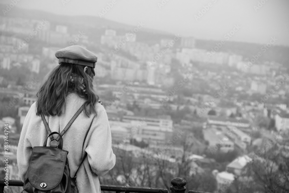 A lonely girl with a leather backpack in a cap is standing on a high mountain and watching the city white and black photo
