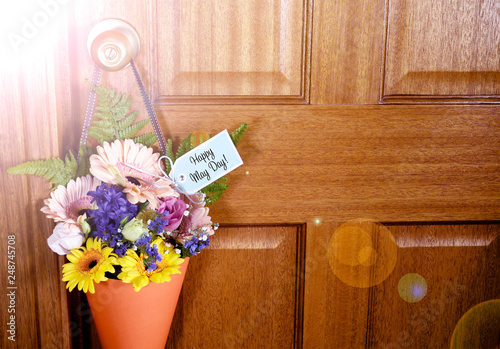 Happy May Day traditional gift of Spring flowers in orange cone hanging from door handle on wooden door, with lens flare.