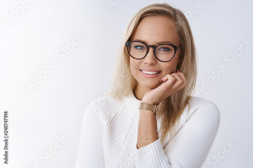 Close-up shot of smart and charming elegant young lady in glasses touching face looking happy and flirty at camera touching face grinning delighted posing against white background