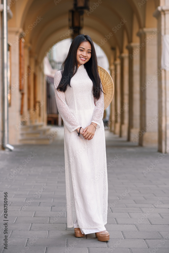 Beautiful happy asian girl dressed in Ao Dai dress with vietnamese conical hat against arches background. Beautiful happy asian girl dressed in Ao Dai dress with vietnamese conical hat against arches background.