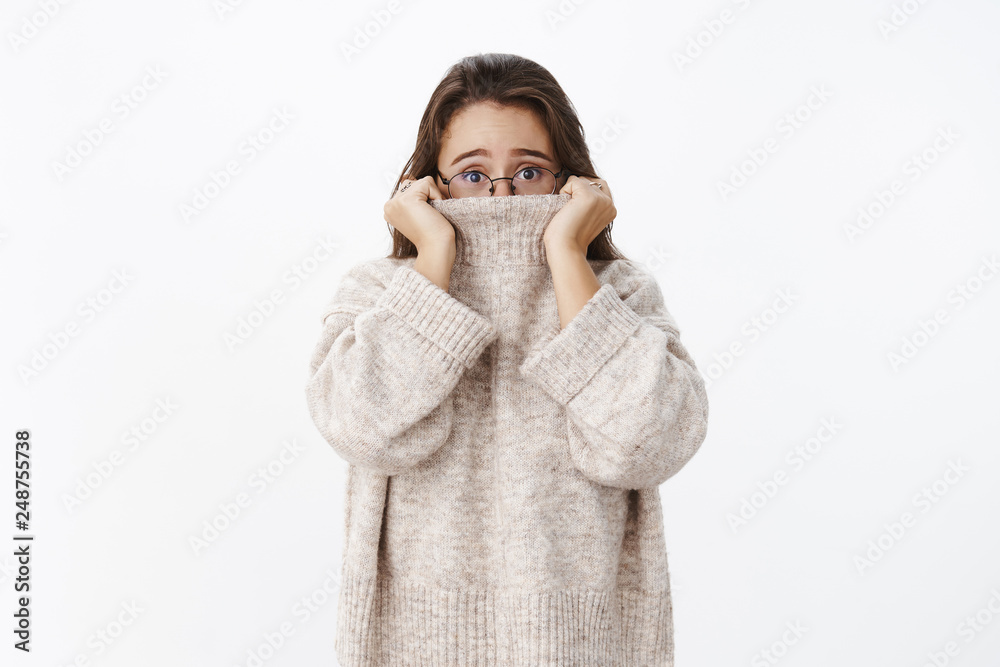 Indoor shot of insecure worried young cute woman in glasses with brown hair pulling collar of sweater on nose and frowning nervously as looking scared at camera, posing anxious over gray background Indoor shot of insecure worried young cute woman in glasses with brown hair pulling collar of sweater on nose and frowning nervously as looking scared at camera, posing anxious over gray background