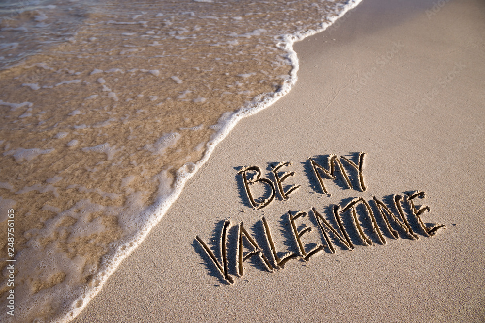 Fototapeta premium Be my Valentine message handwritten on a smooth sand beach with an oncoming wave