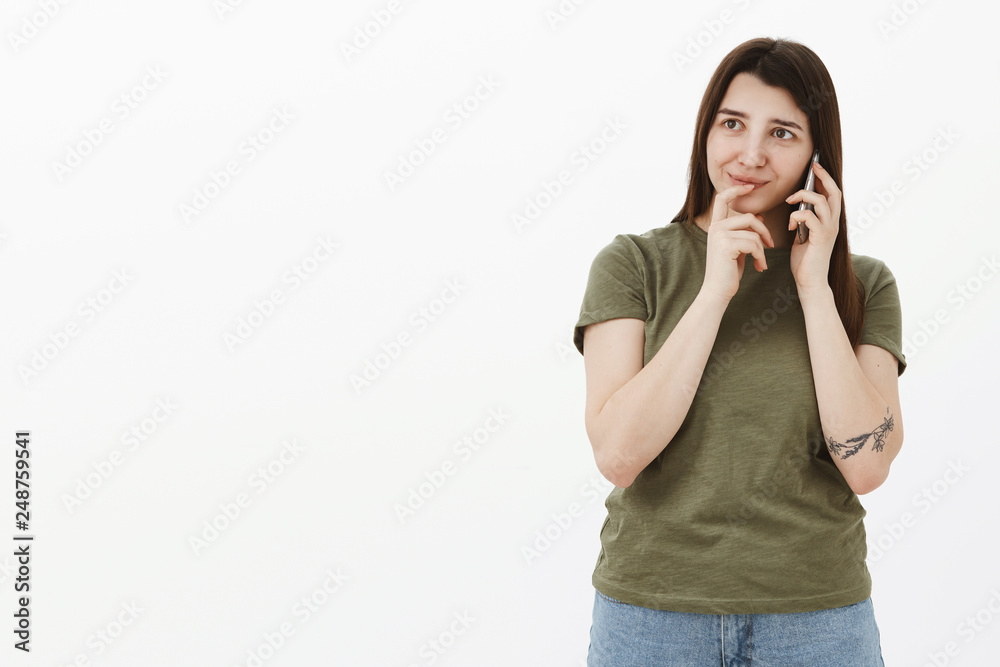 Woman deciding what order, talking with administrator on phone, holding finger on lips as picking making choice or reservation standing amused and thoughtful holding smartphone near ear over gray wall Woman deciding what order, talking with administrator on phone, holding finger on lips as picking making choice or reservation standing amused and thoughtful holding smartphone near ear over gray wall