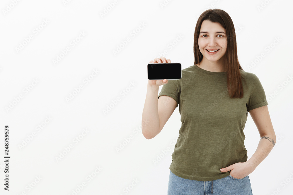 Portrait of friendly and happy young pleased positive brunette in casual t-shirt smiling and showing smartphone in horizontal position as presenting app or mobile phone over gray background Portrait of friendly and happy young pleased positive brunette in casual t-shirt smiling and showing smartphone in horizontal position as presenting app or mobile phone over gray background