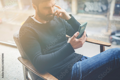 Cheerful bearded hispanic man in black pullover using mobile phone application for mobile blogging. Confident young hipster guy read news from social network at modern loft.Blurred background.Flares