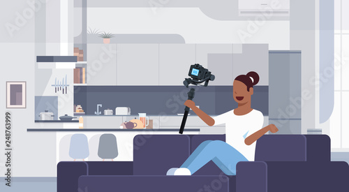 girl blogger recording video on camera african american woman sitting on couch modern kitchen interior using gimbal stabilizer social media blog concept full length flat horizontal