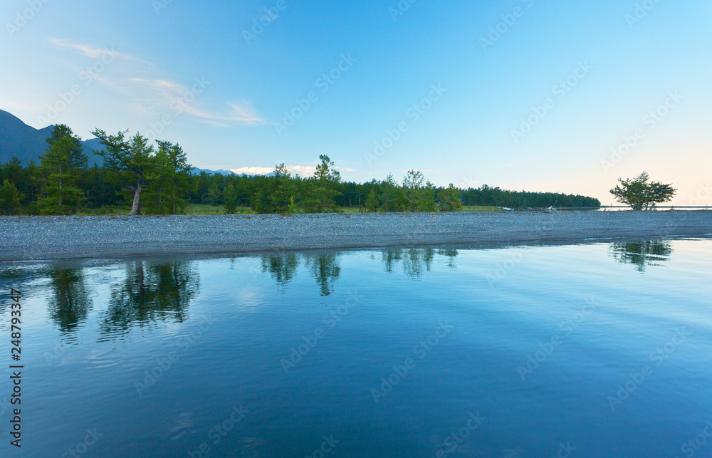 Baikal  Lake on a quiet summer evening. View from the water on the beautiful shore with pebbles  beach near coastal forest.  Natural background