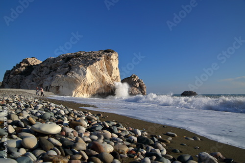 Skała Afrodyty na Cyprze, kamienista piękna plaża omywana falami Morza Śróziemnego, para turystów spaceruje, piękne czyste lazurowe niebo