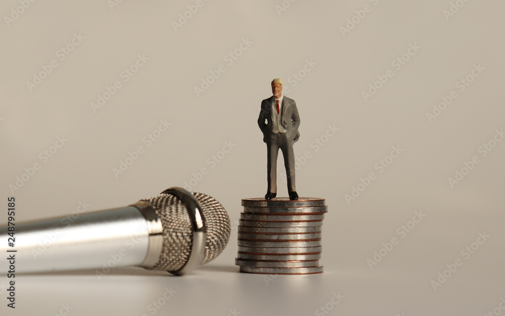 A miniature man standing on a pile of coins next to a microphone. A miniature man standing on a pile of coins next to a microphone.