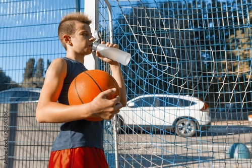 Teenager boy playing street basketball with ball on outdoor city basketball court drinking water photo